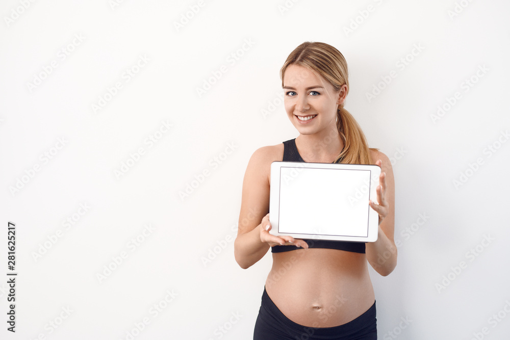 Smiling friendly young pregnant woman holding up a blank white modern tablet with screen to the viewer over a studio background with copy space Smiling friendly young pregnant woman holding up a blank white modern tablet with screen to the viewer over a studio background with copy space