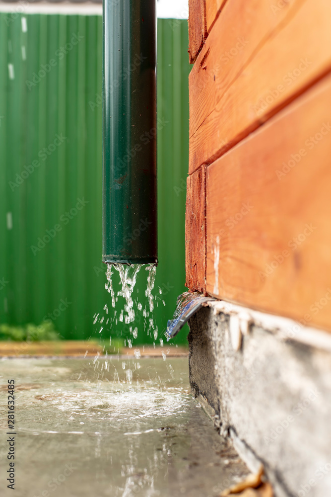 Rain water flows from a drainpipe to a concrete foundation, a wooden