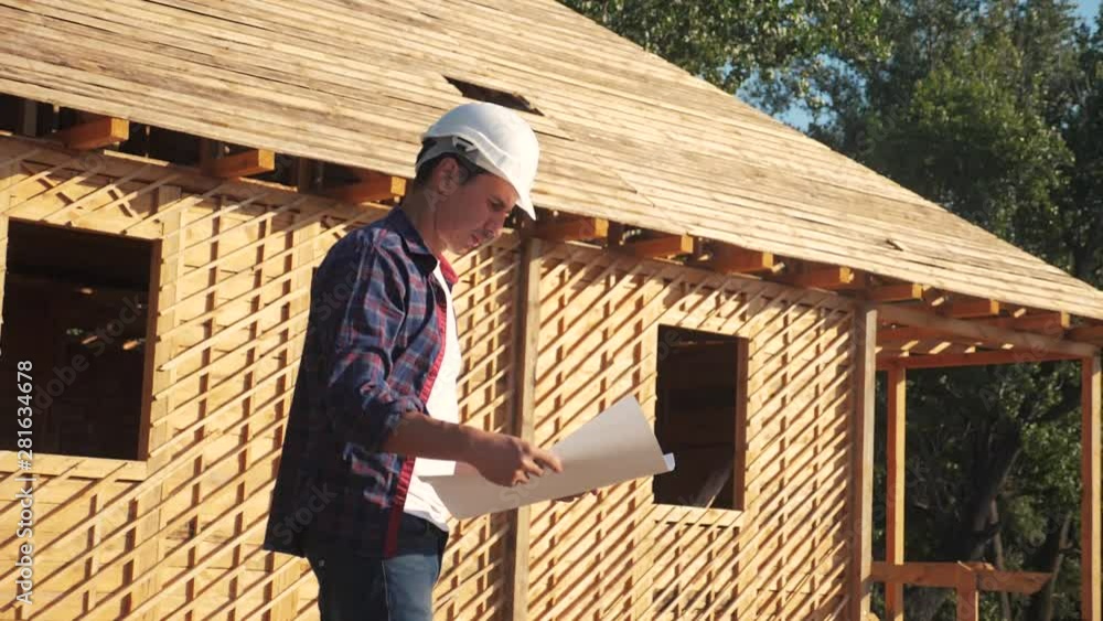 concept building constructing architect slow motion video. man builder in a helmet stands at construction holding a scheme house plan. site near a wooden frame house under construction. worker works concept building constructing architect slow motion video. man builder in a helmet stands at construction holding a scheme house plan. site near a wooden frame house under construction. worker works