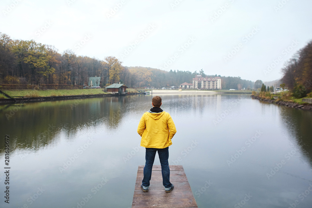 Stylish young man in yellow waterproof raincoat standing on the wooden pier by the lake in the park at autumn. Back view.