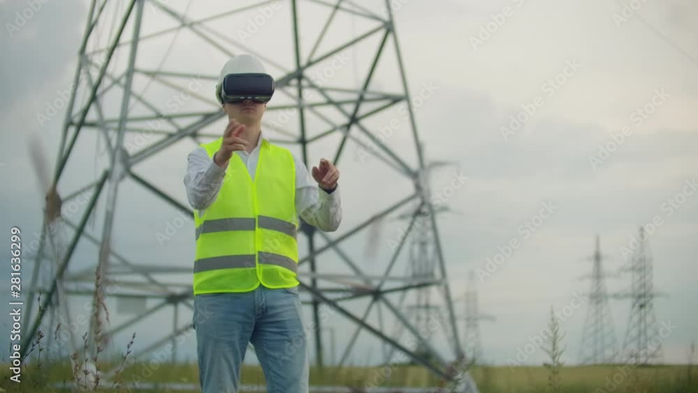 A male electrician in virtual reality glasses moves his hand simulating the work with the graphical interface of the power plant against the background of high-voltage electric transmission lines A male electrician in virtual reality glasses moves his hand simulating the work with the graphical interface of the power plant against the background of high-voltage electric transmission lines