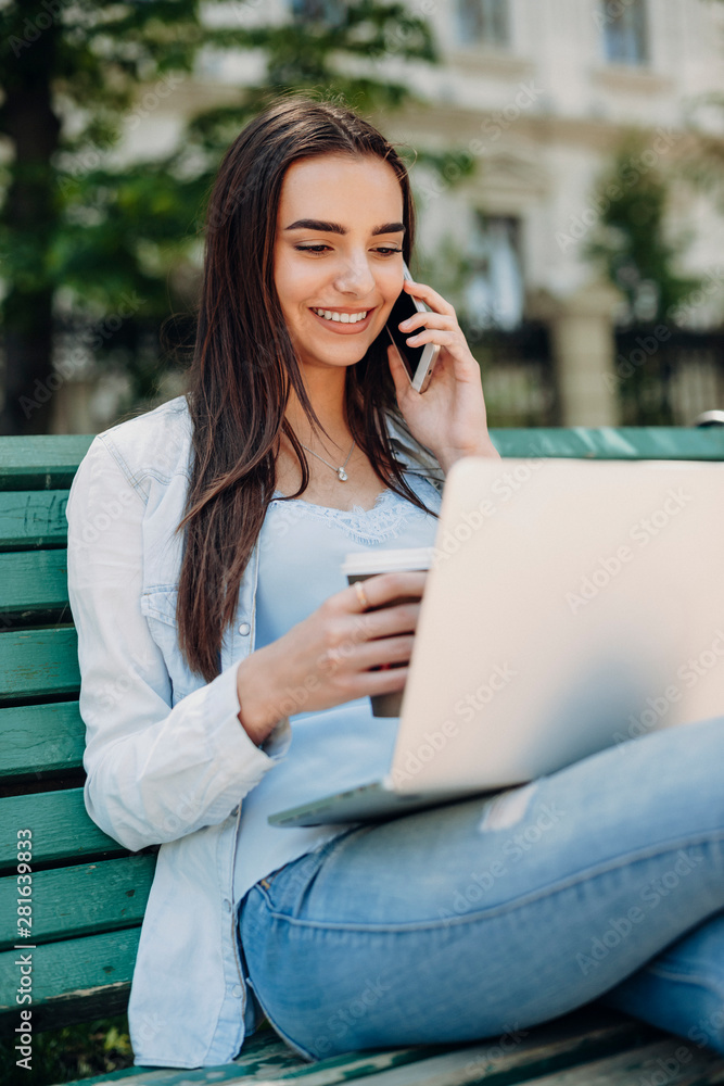 Lovely young female talking at her smartphone while looking at screen of a laptop while sitting on a bench and drinking coffee outside. Lovely young female talking at her smartphone while looking at screen of a laptop while sitting on a bench and drinking coffee outside.