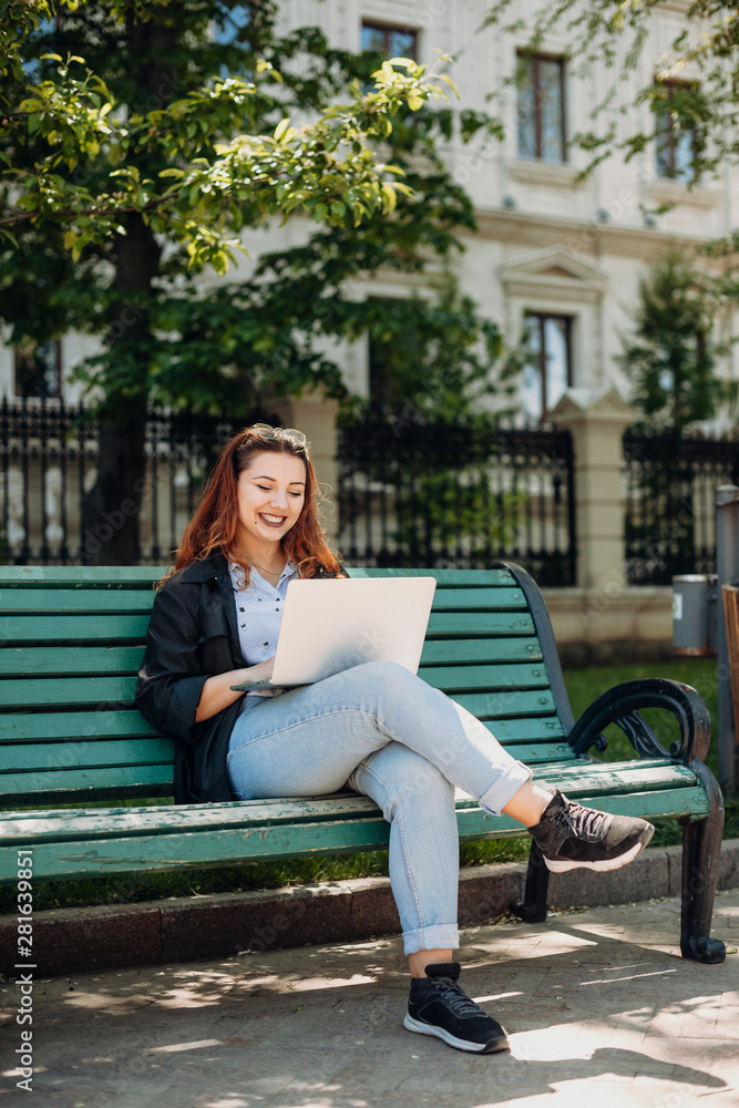 Portrait of a cheerful plus size women with long red hair sitting on a bench smiling with a laptop on her legs outside in the park working. Portrait of a cheerful plus size women with long red hair sitting on a bench smiling with a laptop on her legs outside in the park working.