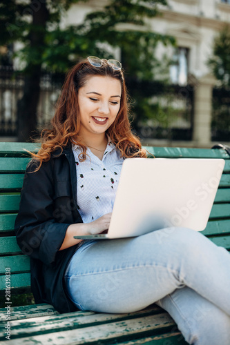 Portrait of a cherful pluss size woman with long red hair siting on a bench smiling with a laptopt on her legs outide in the park working. photo