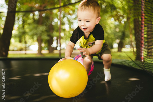 happy and smiling two-year-old boy playing with two balls on a trampoline in the park in summer while walking with his family