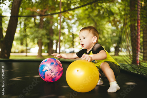 happy and smiling two-year-old boy playing with two balls on a trampoline in the park in summer while walking with his family
