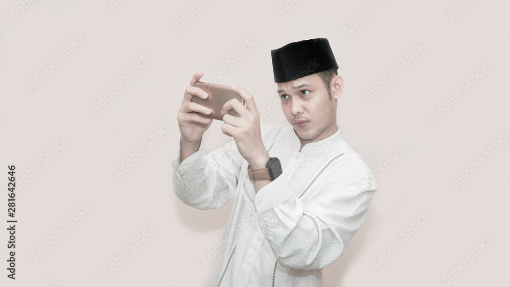 portrait of handsome asian muslim man with head cap and using smartphone and seriously playing video game portrait of handsome asian muslim man with head cap and using smartphone and seriously playing video game