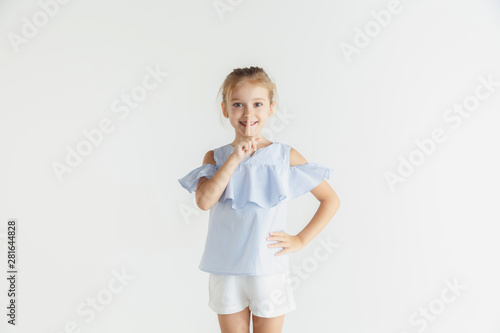 Stylish little smiling girl posing in casual clothes isolated on white studio background. Caucasian blonde female model. Human emotions, facial expression, childhood, sales concept. Whispering a