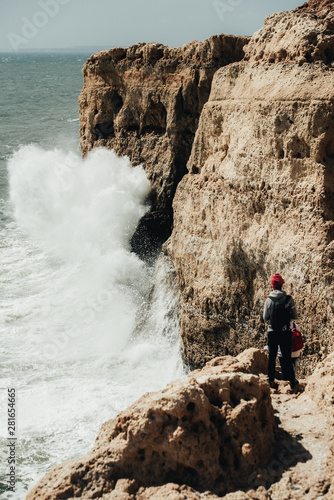 Young tourist standing on the rim of cliff. Man enjoying awesome view on the boulders on the rocky seashore. Attractive boy in black eyeglasses on ocean wave background. Mountain summer landscape