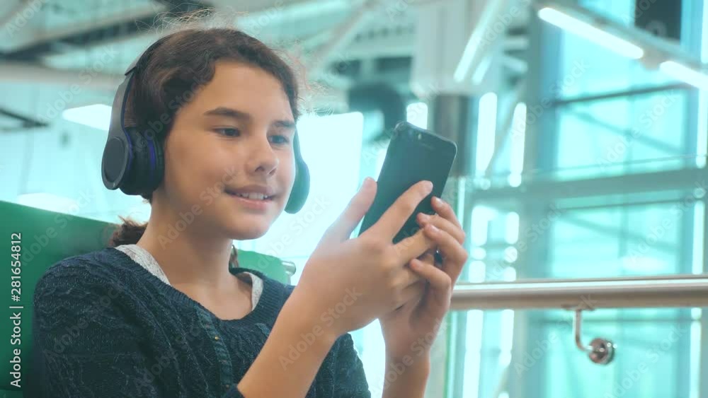 airport waiting hall room for a flight by plane. young happy teen girl in headphones listening to the music lifestyle on smartphone chatting communicates in the messenger. teenager girl in social airport waiting hall room for a flight by plane. young happy teen girl in headphones listening to the music lifestyle on smartphone chatting communicates in the messenger. teenager girl in social
