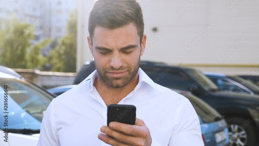 Young Attractive Man Standing on Parking near Office Building Typing a Message on his Smartphone. Handsome Businessman in White Shirt Using Smartphone outdoor. Young Attractive Man Standing on Parking near Office Building Typing a Message on his Smartphone. Handsome Businessman in White Shirt Using Smartphone outdoor.