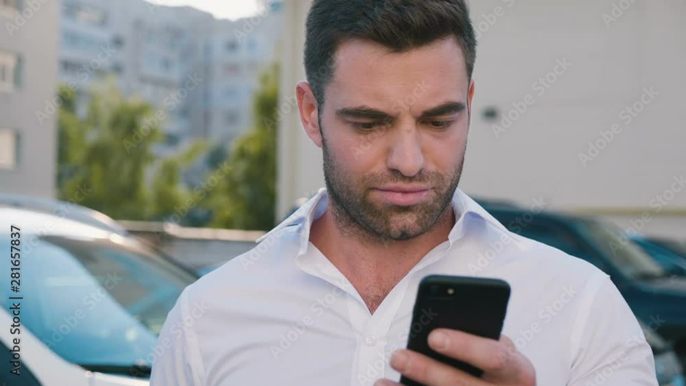 Young Attractive Man Standing on Parking near Office Building Typing a Message on his Smartphone. Handsome Businessman in White Shirt Using Smartphone outdoor. Young Attractive Man Standing on Parking near Office Building Typing a Message on his Smartphone. Handsome Businessman in White Shirt Using Smartphone outdoor.