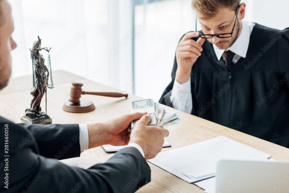 cropped view of man holding dollar banknotes near judge in glasses gesturing in office cropped view of man holding dollar banknotes near judge in glasses gesturing in office