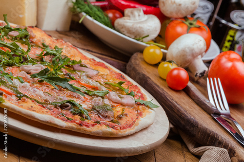 Italian pizza with ham, tomatoes, cheese and arugula on a wooden board, the background is wooden decorated with vegetables and cutlery