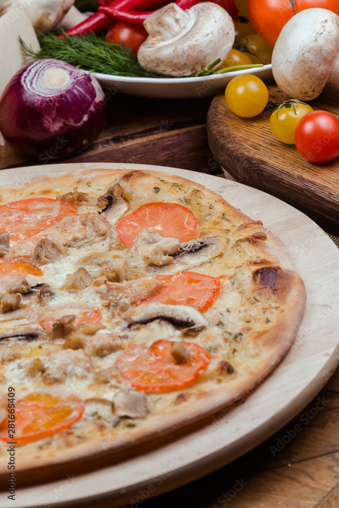 Italian pizza with chicken, tomatoes, mushrooms and cheese on a wooden board, the background is wooden decorated with vegetables and cutlery