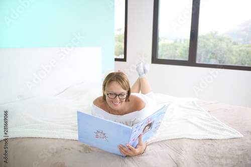 Happy woman viewing a family photo album lying face down and wearing glasses on the bed in the bedroom in an environment corresponding to an scandinavian concept called Lagom