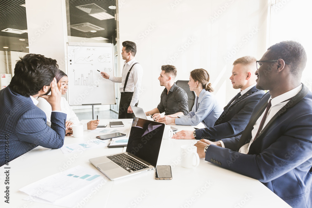 Obraz premium Company leader standing near the flipchart. A team of young businessmen working and communicating together in an office. Corporate businessteam and manager in a meeting.