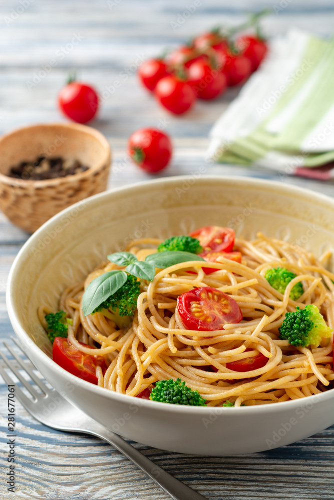 Whole wheat spaghetti pasta with broccoli, cherry tomatoes and basil in bowl on wooden background. Selective focus.