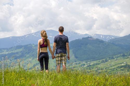 Young couple, sportive man and slim woman holding hands outdoors on background of beautiful mountain landscape on sunny summer day, back view.