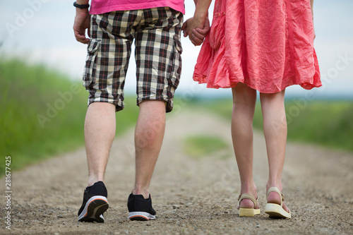 Back view of legs of young slim woman in red dress and man in shorts walking together by ground road on sunny summer day on blurred background.