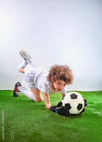 Goalkeeper lying on the grass catches a ball. Excited little toddler boy playing football on soccer field against light background. Active childhood and sports passion concept. Save space