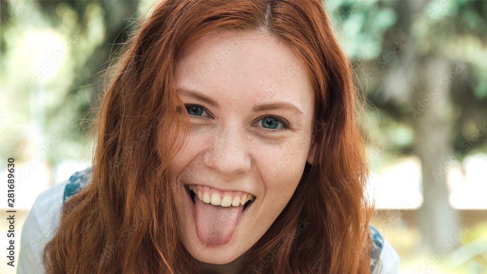 Red-haired girl making faces. Freckled ginger girl walks in the park close-up. Young red-headed woman on blurred background outdoor.