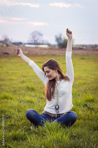 Beautiful young girl sitting in the grass, closed eyes, hands above her head.