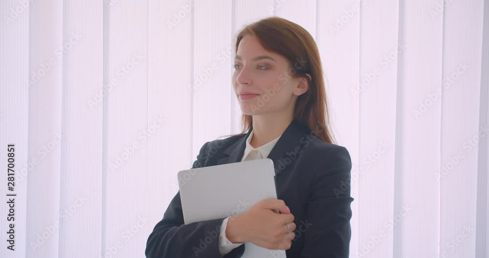 Closeup portrait of young caucasian businesswoman holding the laptop looking at camera standing indoors in the white office Closeup portrait of young caucasian businesswoman holding the laptop looking at camera standing indoors in the white office