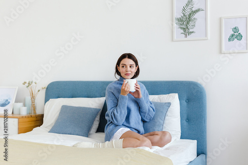 Girl in a blue sweater in interior Hygge style with a cup in hand sits on the bed