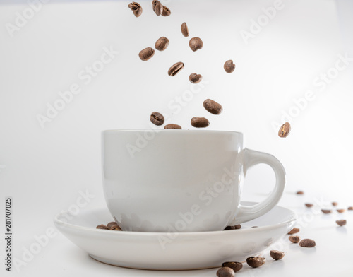 Coffee beans fall into a white porcelain cup and saucer, bouncing and jumping through the air conveying alert excitement, all isolated against a white background