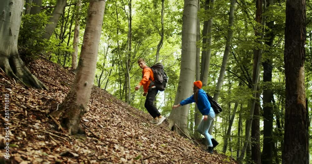 Male tourist with backpack supporting girlfriend while climbing on woody hill, hiking in mountains, recreation