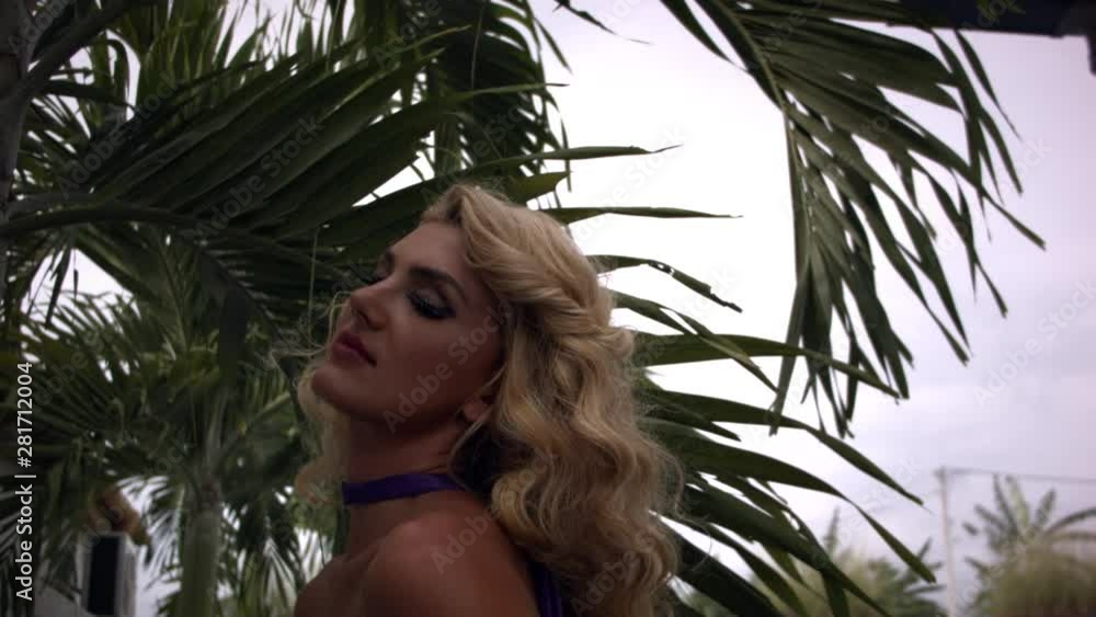 Close up view of young pretty woman with blonde curly hair in purple dress posing and looking in camera on background of palm leaves. Close up view of young pretty woman with blonde curly hair in purple dress posing and looking in camera on background of palm leaves.