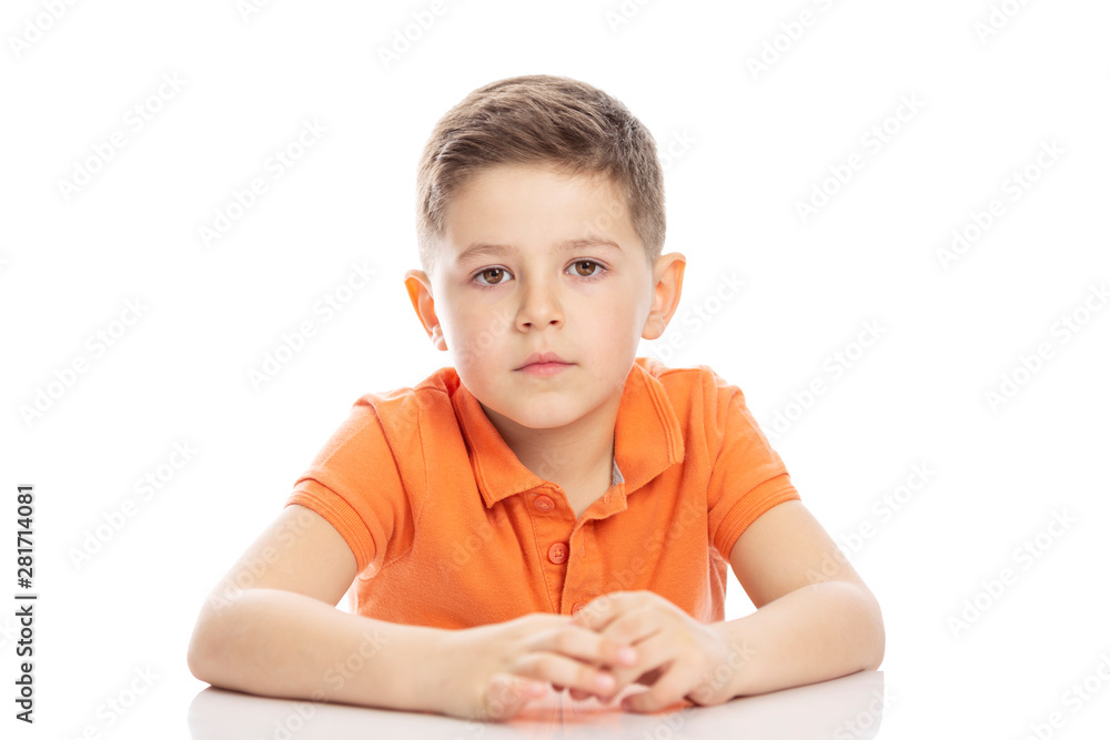 A serious school-age boy in a bright orange polo t-shirt is sitting at the table. Close-up. Isolirvoan on a white background. A serious school-age boy in a bright orange polo t-shirt is sitting at the table. Close-up. Isolirvoan on a white background.