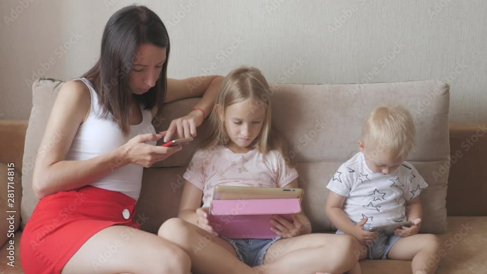 Young mother, daughter and little son are using gadgets lying on a white bed. Concept modern family during weekend at home. Contemporary technology. Young mother, daughter and little son are using gadgets lying on a white bed. Concept modern family during weekend at home. Contemporary technology.