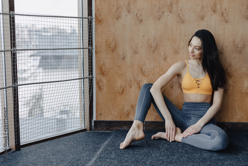 Young attractive fitness girl sitting on the floor near the window on the background of a wooden wall, resting on yoga classes Young attractive fitness girl sitting on the floor near the window on the background of a wooden wall, resting on yoga classes