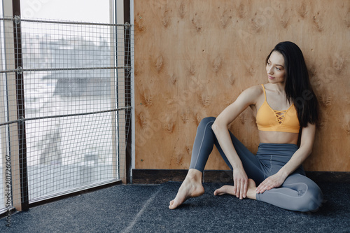 Young attractive fitness girl sitting on the floor near the window on the background of a wooden wall, resting on yoga classes © pressahotkey