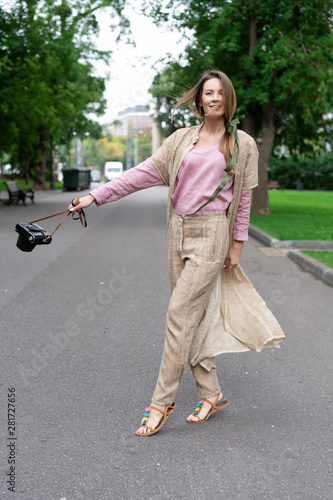young European girl, natural appearance, long hair, beautiful face, coffee colored trousers and jacket, pink shirt is fun to walk on pavement city park. Comfortable linen womens clothing.