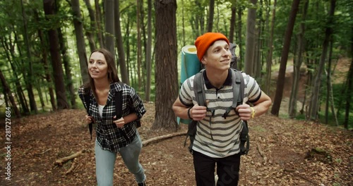 Handsome male and pretty female hikers with backpacks walking uphill in forest, chatting and smiling, adventurers