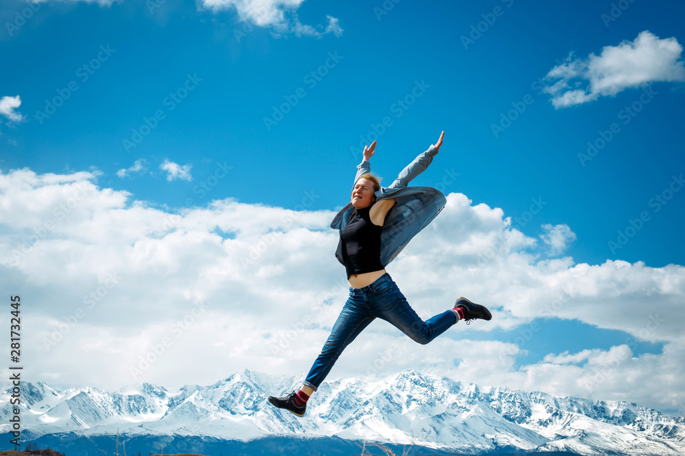 Young woman in jeans jumps high into the air on the background of mountain peaks covered with snow and blue sky with white clouds. Spectacular views of mountain range. Travel and discovery concept. Young woman in jeans jumps high into the air on the background of mountain peaks covered with snow and blue sky with white clouds. Spectacular views of mountain range. Travel and discovery concept.