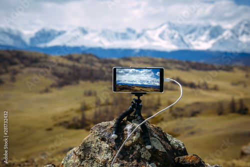 Smartphone on tripod mounted on stone in front of  stunning view of the snow-capped peaks. Image is focused on display. Gadget records timelapse of moving of clouds over the North Chui mountain range. © exebiche