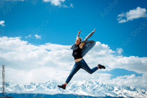 Young woman in jeans jumps high into the air on the background of mountain peaks covered with snow and blue sky with white clouds. Spectacular views of mountain range. Travel and discovery concept.