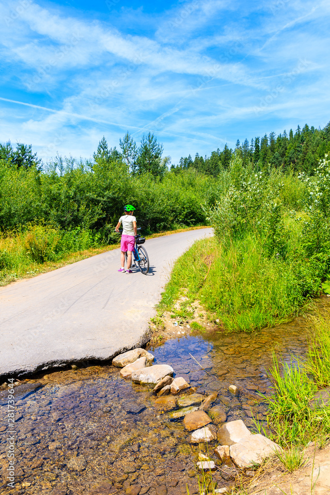 Young woman cyclist standing with bike on road near small stream by Dunajec river, Nowy Targ, Tatra Mountains, Poland