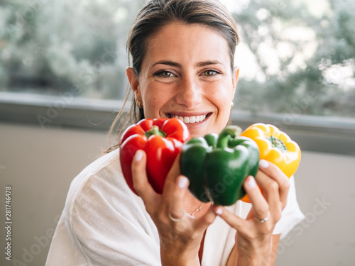 Caucasian girl sitting in her kitchen showing three big colored peppers. Green, red and yellow