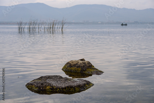 The water expanses of the endless Lake Sevan with stones in the foreground grass and two fishermen on a cloudy day with mountain views in the background.