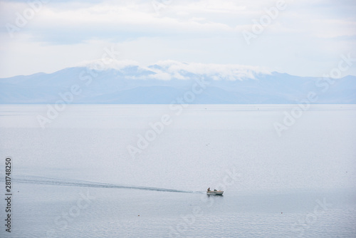 The water expanses of the endless Lake Sevan with stones in the foreground grass and two fishermen on a cloudy day with mountain views in the background.