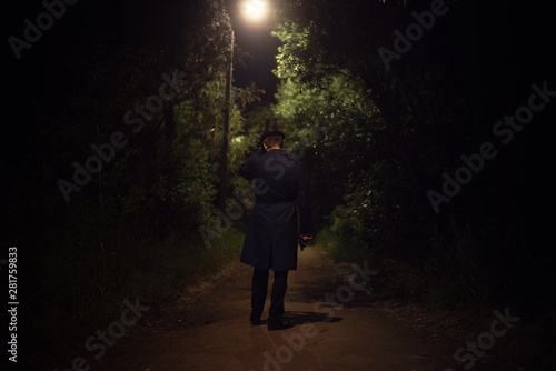 Detective agent in a leather hat and coat is walking through the night park with a handgun in a hand.