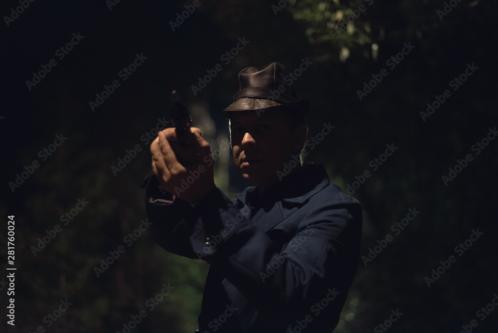 Thrilled Detective agent in a leather hat and coat is standing and aiming into the dark by a gun.