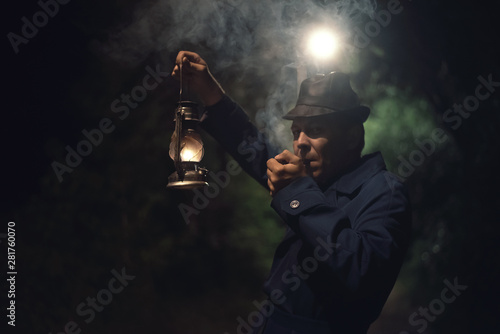 Quadro em tela Detective agent in a hat and coat is holding in hand a kerosene lamp and smokes his smoking pipe