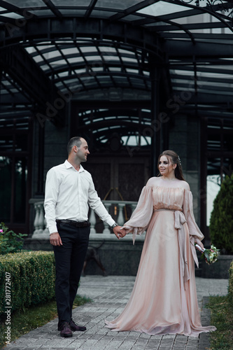 Outdoor shot of young couple in love walking near house. Man and woman. Couple in love: young girl in a long luxury dress and young boy in in black trousers and a white shirt. Couple in love