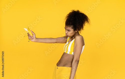 Funny dangerous african american teen girl pretending holding banana gun pistol looking aiming, silly cool black young ethnic woman make shooting gesture isolated on summer yellow studio background